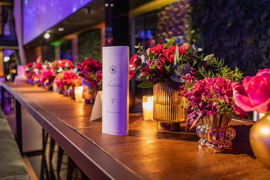 A long wooden table decorated with a series of floral arrangements featuring pink and purple flowers, including roses and hydrangeas, in gold and amber glass vases. The flowers are fresh, with vibrant