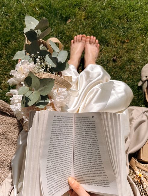 A person seated outdoors on a grassy area, holding an open book with dense text across two pages. Next to them, there is a bouquet of fresh flowers featuring white roses, small white filler flowers, a
