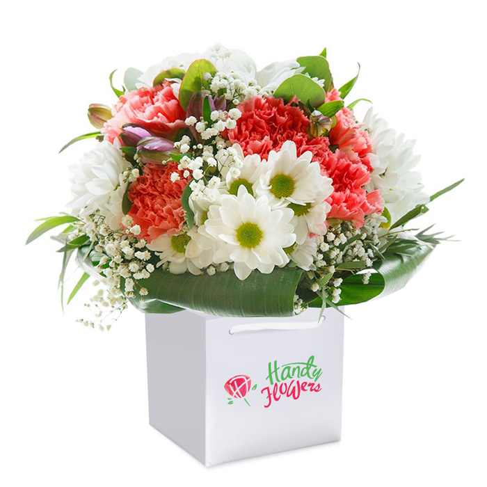 Resting gracefully within a crisp white gift bag, this radiant bouquet is a celebration of Upper Clapton's blossoming spirit and refined florist skill. At its heart are abundant coral and peach hued carnations, each petal layered in ruffles that evoke a sense of softness and perennial renewal-like spring evenings along the canal. These are offset by striking white daisies, their bold lime-green centers glowing with positivity, creating a vibrant counterpoint that brightens the whole arrangement. Scattered with intent, dainty sprays of pure white baby's breath add a dreamy, ethereal texture, softening transitions between the more prominent blooms. Subtle purple-tipped buds hide within the flowers, adding dimension and the subtle excitement of new beginnings, reminiscent of freshly opened markets on Clapton's high streets. Encircling everything is a lush and glossy ring of deep green foliage, skillfully shaped to support and accentuate the blooms, its lustre catching the gentle light much like leaves after early summer rain. The arrangement is composed in a perfectly rounded mound, evidence of careful attention and a florist's practiced touch. The sturdy white gift bag, designed for easy delivery and adorned with simple ribbon handles, enhances the sense of occasion, while the pure white setting highlights every colourful detail. Crafted by Florist Upper Clapton, this bouquet captures the joy of celebration-be it for birthdays, anniversaries, or spontaneous gestures of kindness-delivered fresh across the local neighbourhood.