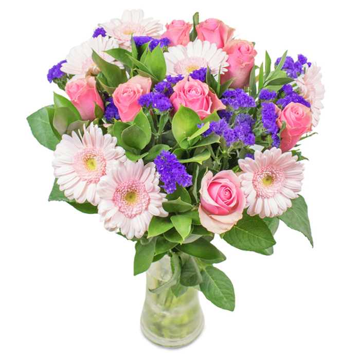A vibrant, artistically arranged floral bouquet stands at the heart of the image, nestled in a clear glass vase on a pristine surface that evokes the simplicity of Upper Clapton's neighbourhood interiors. The arrangement is anchored by approximately seven to nine classic pink roses, their petals in varying stages from tightly furled buds to gentle opening spirals, displaying a graceful progression that recalls the tranquil rhythm of life along the River Lea. Intertwined among them are five to seven pale pink gerbera daisies, their large, rounded heads and radiating petals glowing softly-a visual echo of spring blossoms in Millfields Park. Striking clusters of deep purple statice thread through the bouquet, offering a textured contrast and bold colour without overwhelming the palette. Rich, deep-green foliage surrounds the blooms, enhancing both the sense of natural abundance and the arrangement's freshness, like a recently gathered display from a local Saturday flower market. The transparent vase allows glimpses of elegantly arranged stems and sparkling water, making the bouquet appear fresh-cut-a thoughtful detail that speaks to the high standards of flower delivery and care in Upper Clapton. Designed to suit a wide array of occasions-from birthdays and anniversaries to a welcoming gift for a new neighbour in Clapton Square-this local creation radiates celebration, warmth, and heartfelt affection.